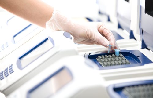 A gloved hand is placing a sample tube into a thermal cycler among several other machines.