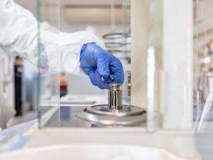 A gloved hand adjusts a calibration weight on a balance scale in a laboratory setting.