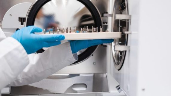 A technician in a lab coat inserts a tray with instruments into an autoclave for sterilization.